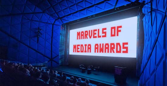 people sit in a darkened theater with a bright image on a big screen that features the words Marvels of Media Awards in red