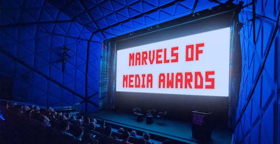 Redstone Theater at Museum of the Moving Image people sit in a darkened theater with a bright image on a big screen that features the words Marvels of Media Awards in red